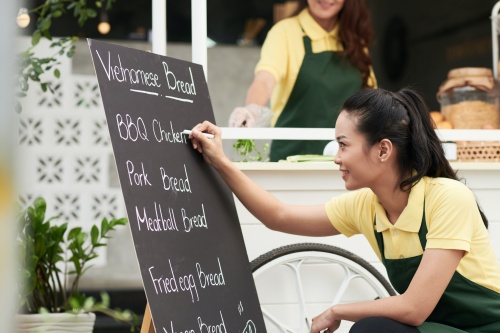 Smiling woman writing menu on board in front of street food cart with piece of white chalk